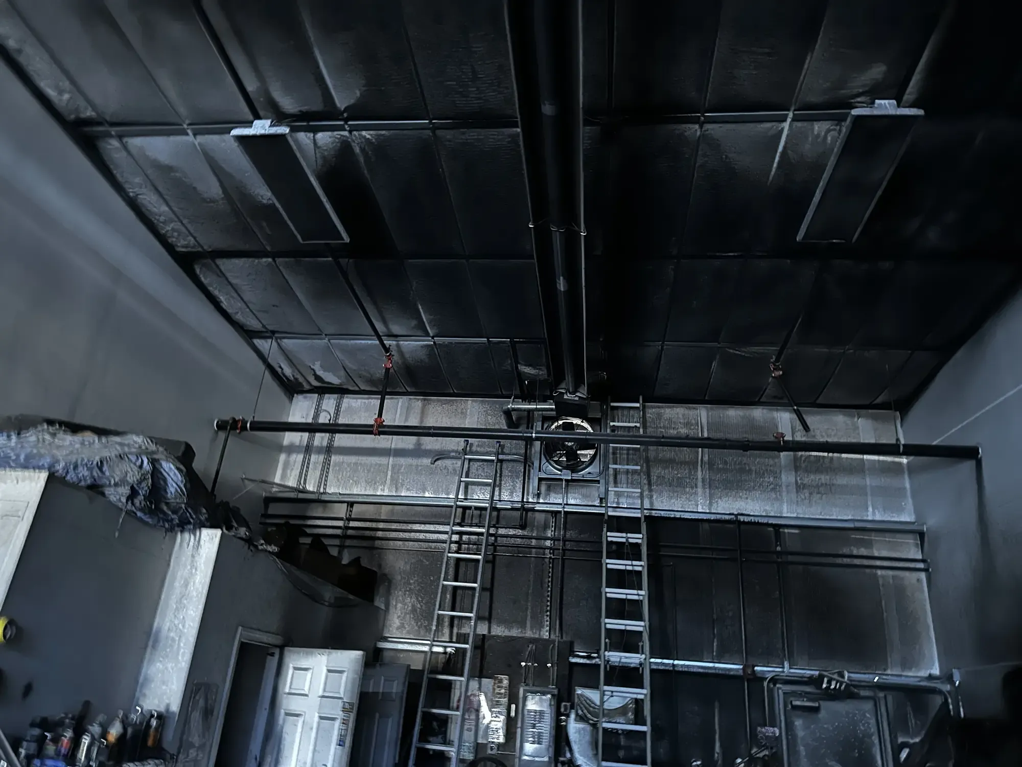A commercial interior after a fire, with a soot-blackened ceiling, charred ductwork, and ladders leaning against the far wall.