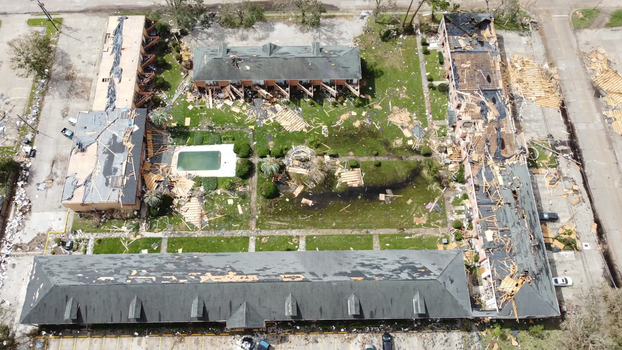 An aerial view of a large multi-building commercial complex with widespread roof damage and debris across the property.
