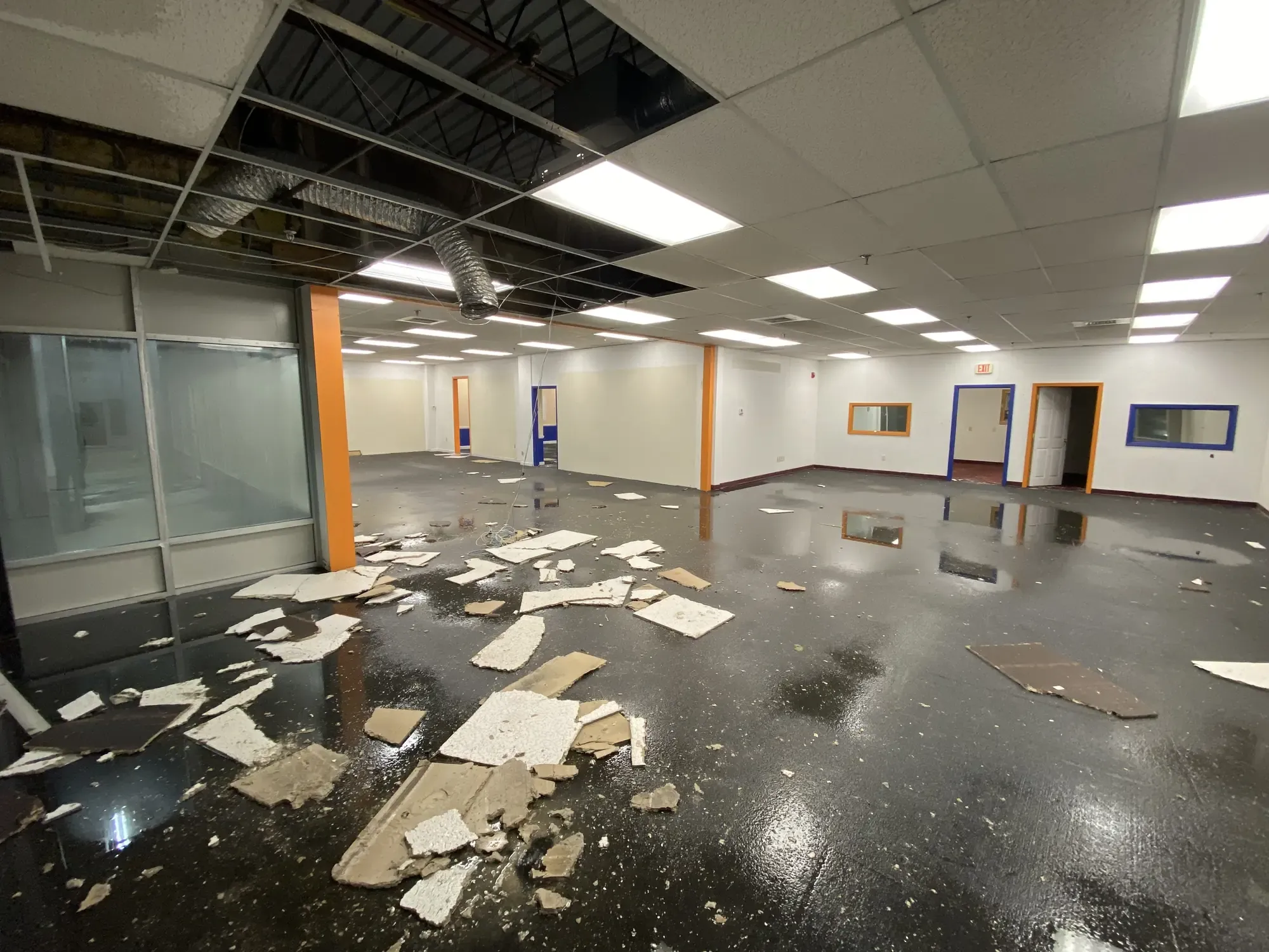 A commercial interior after a water loss, with fallen ceiling tiles scattered across a wet floor and ductwork exposed overhead.