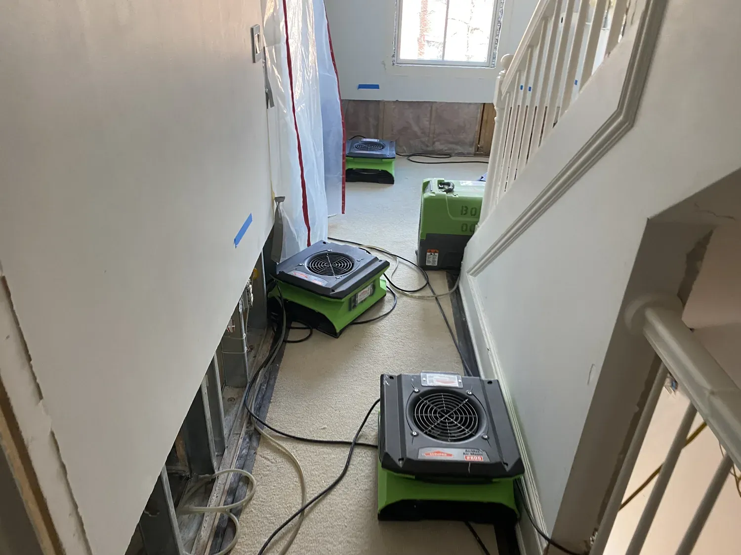 A residential hallway under active mitigation, with air movers and a dehumidifier drying the carpet and stairs.