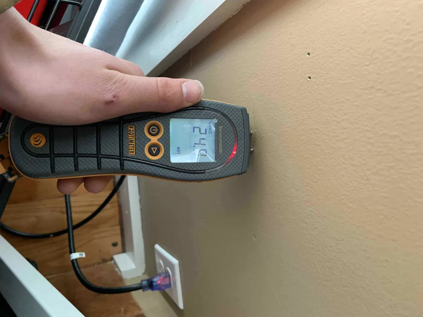 A pin-type moisture meter held against a wall, its display reading a high moisture level marked WET.