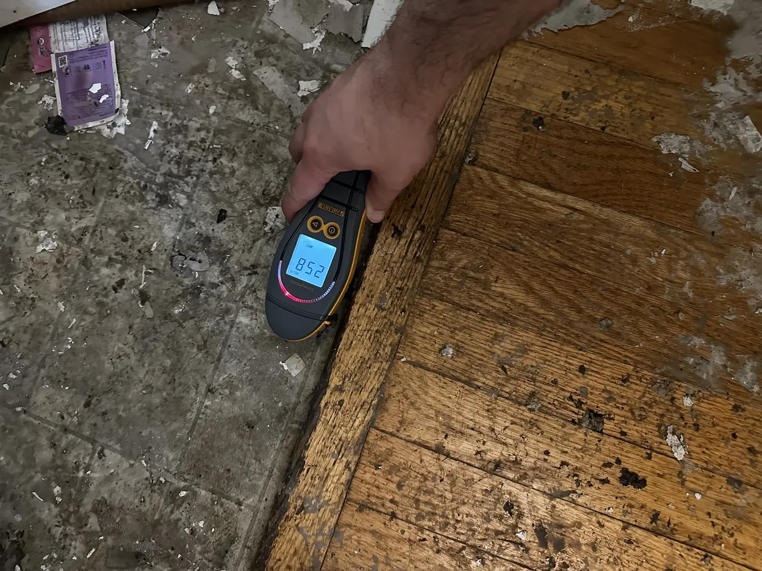 A moisture meter held against a wet hardwood floor during on-site documentation, its display showing a high reading.