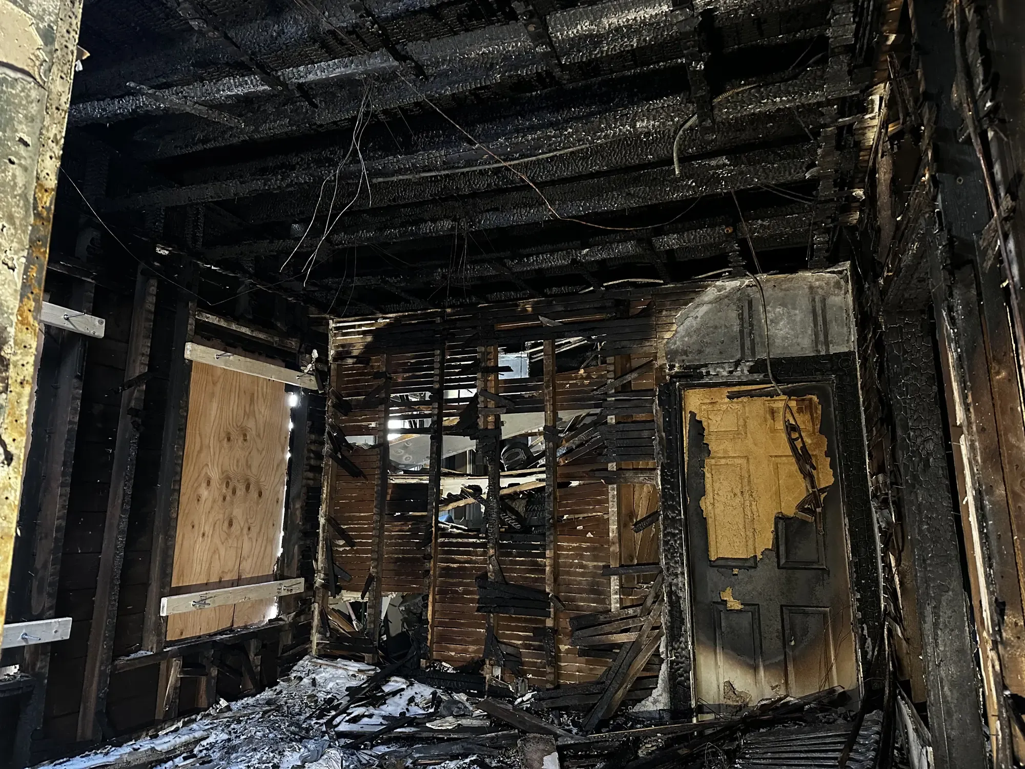 A fire-damaged residential interior burned to charred framing, with a boarded-up window and debris across the floor.