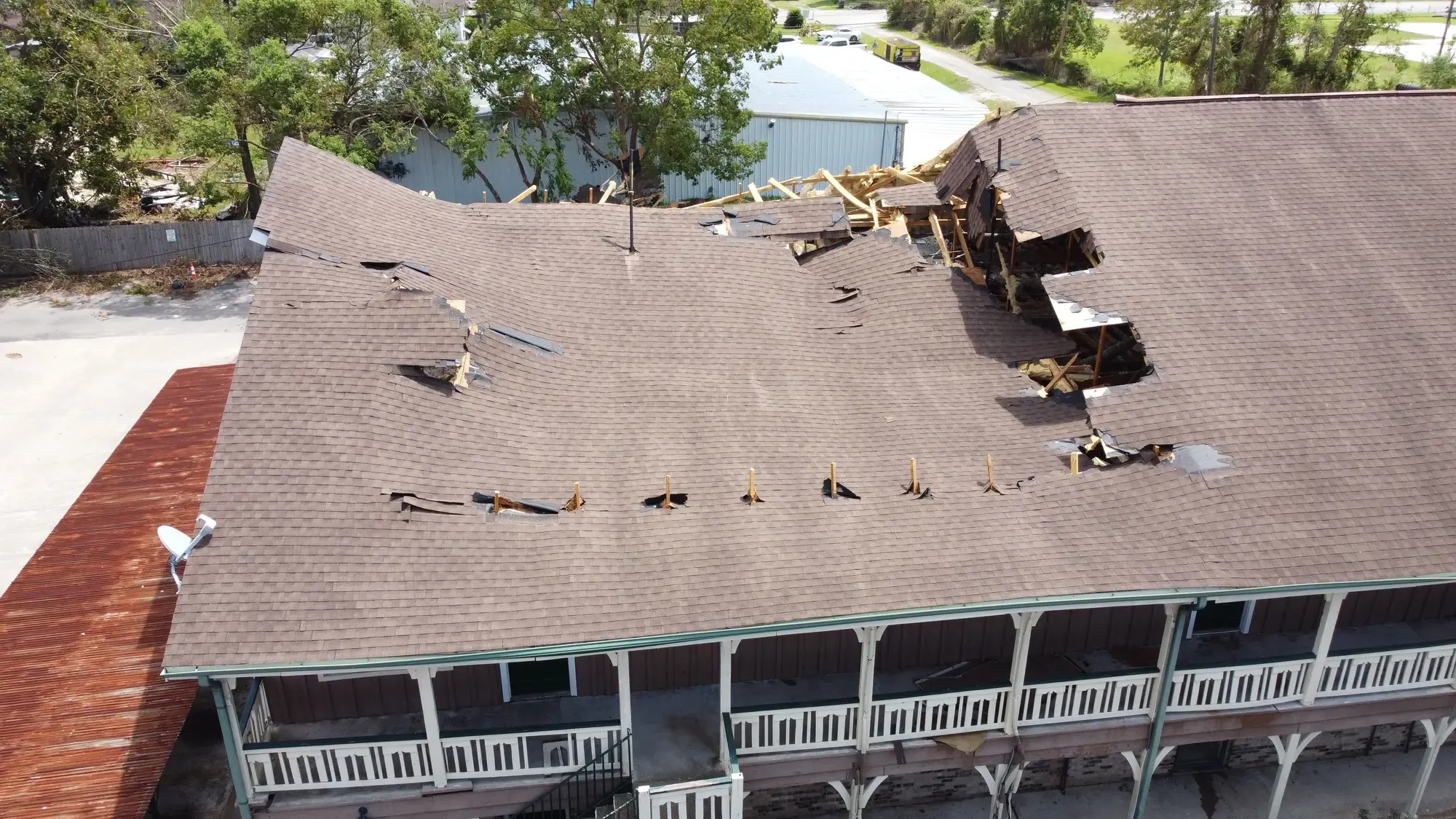 An aerial view of a storm-damaged roof with shingles stripped away and a section of framing torn open to the sky.