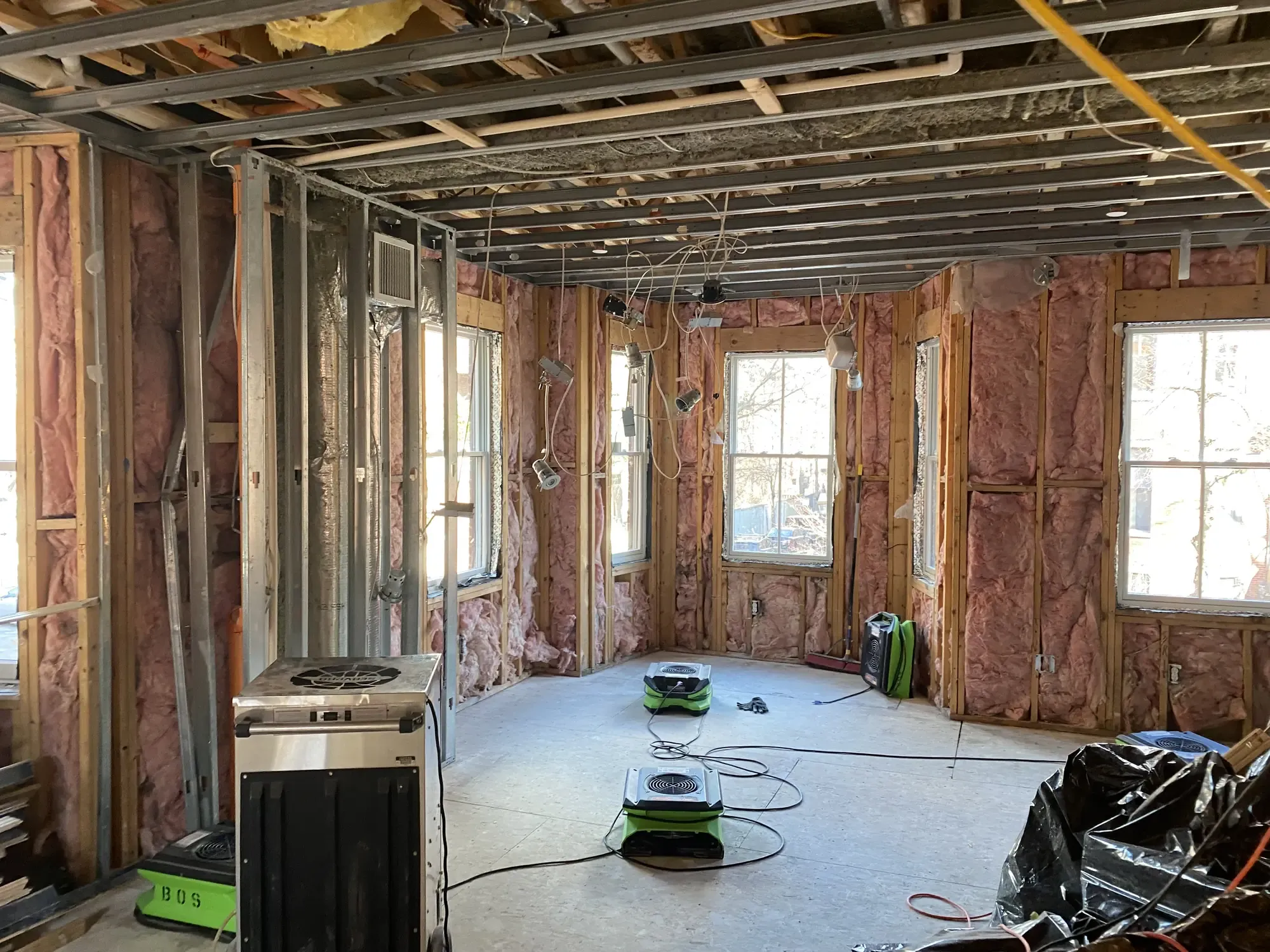 A gutted residential room dried down to the studs, with exposed pink insulation, an open ceiling, and air movers running on the bare subfloor.